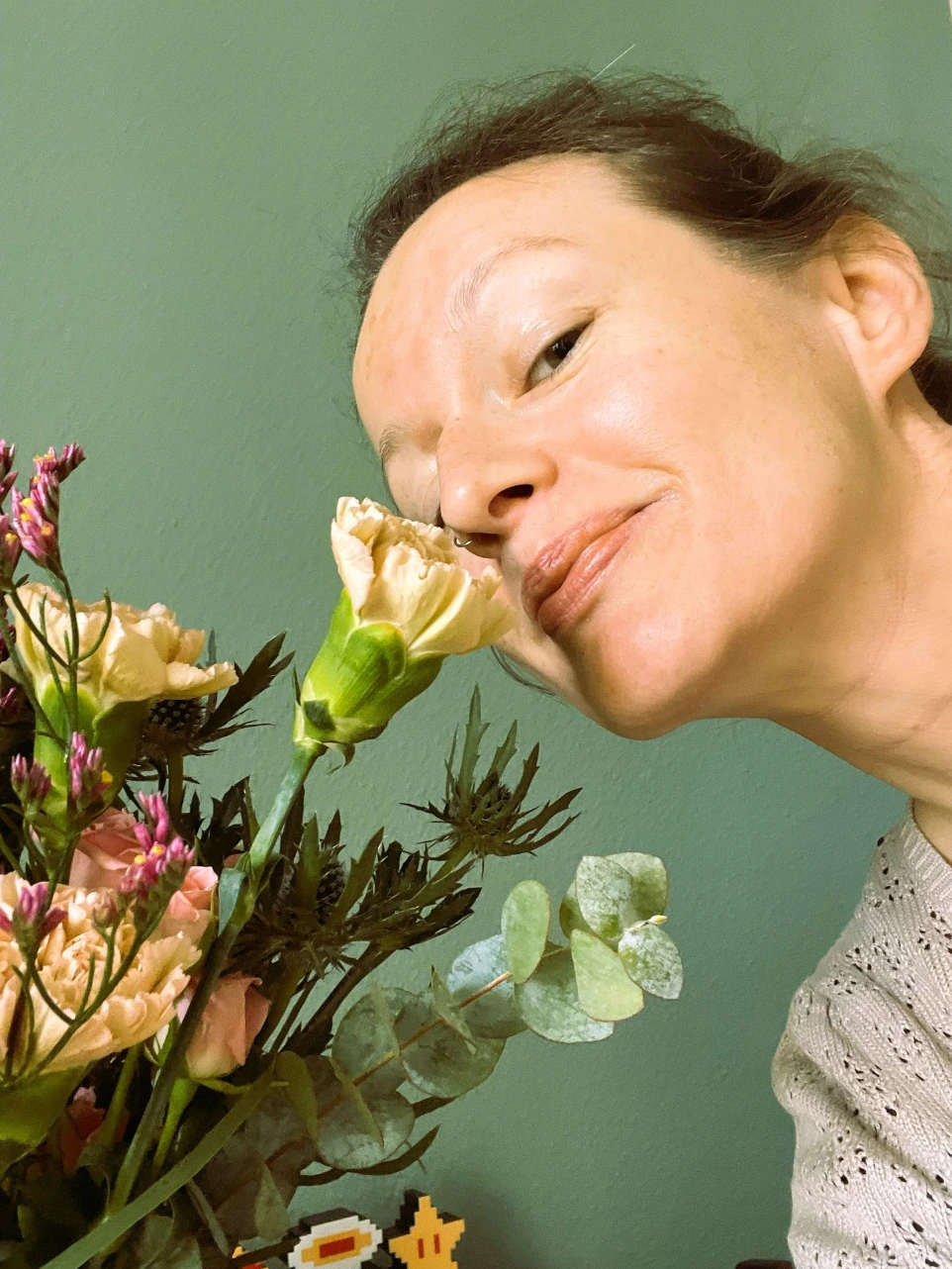 Die Gründerin von Blätterwerk, Therese, riecht an einem pastellfarbenem Blumenstrauß vor einer grünen Wand. Das Bild vermittelt ein Gefühl von Lebensfreude und Naturverbundenheit.